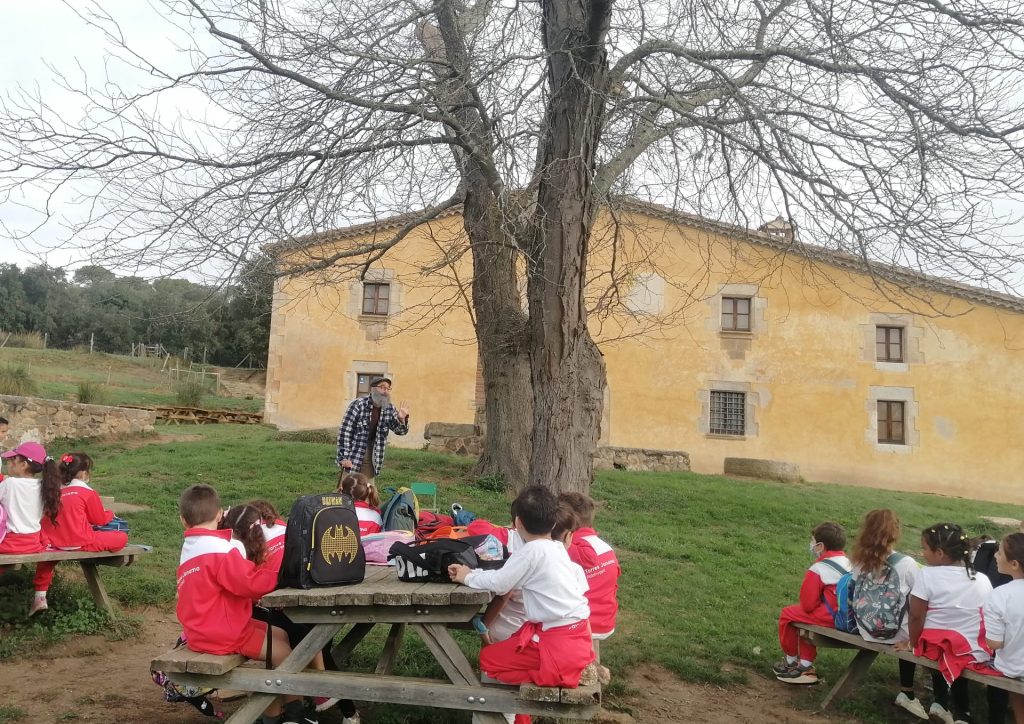 Fotografia de l'activitat d'etnobotànica amb la masia de can Vilallonga al fons i els escolars i educador en primer terme.