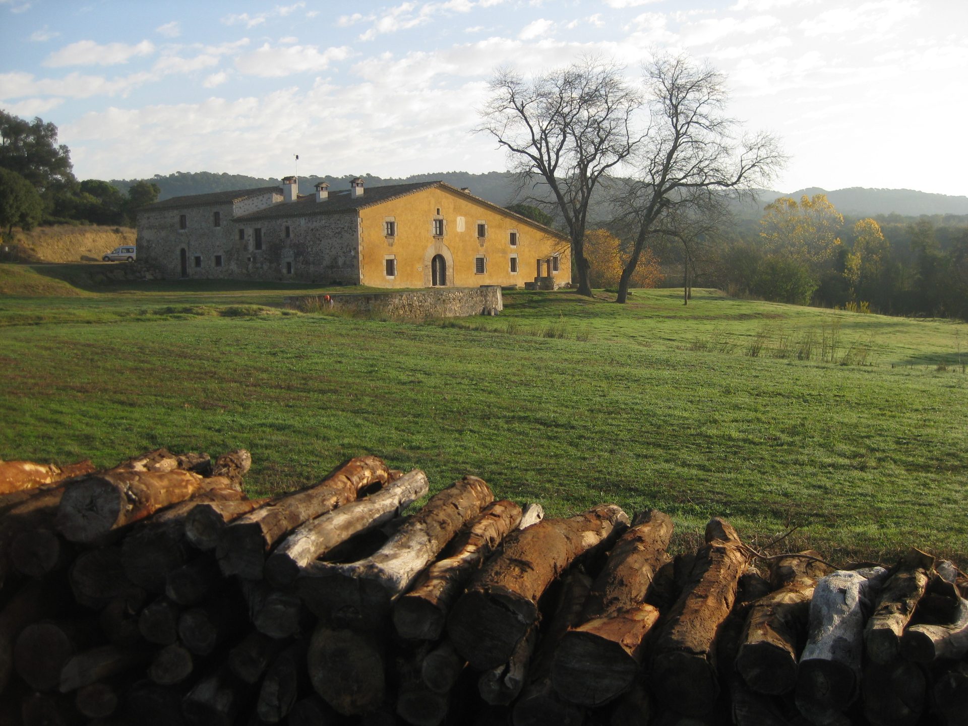Al fons es veu la masia de Can Vilallonga, envoltada de prats i amb una filera de llenya a primer terme, testimoni de l'explotació forestal.